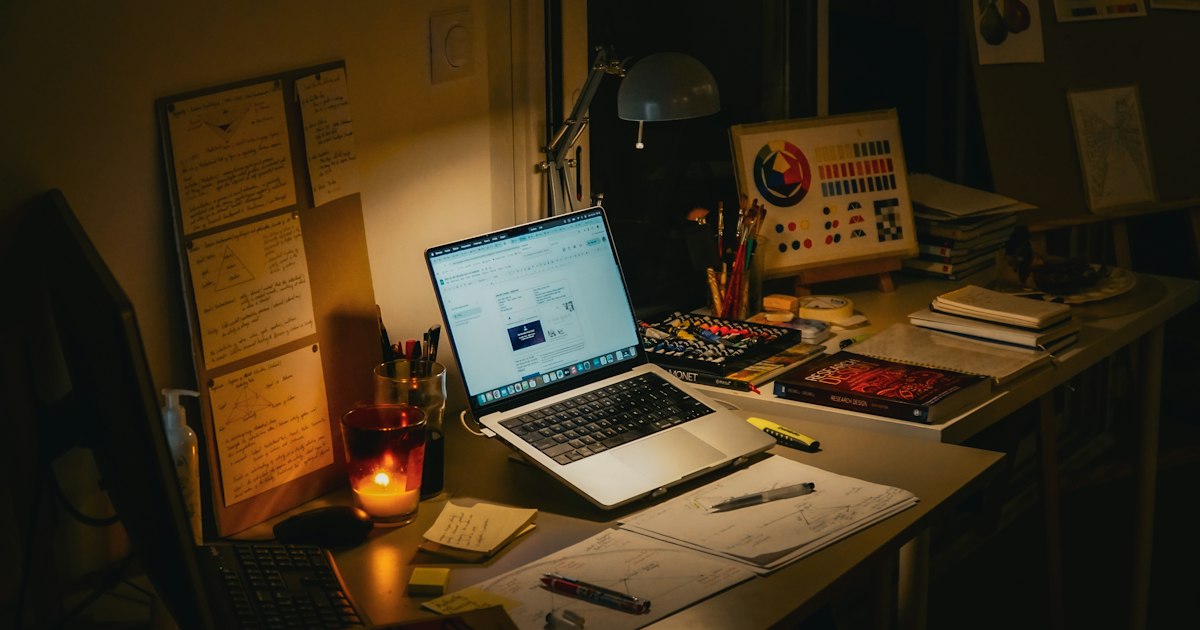 Escritorio de noche con un portátil encendido, una vela y papeles